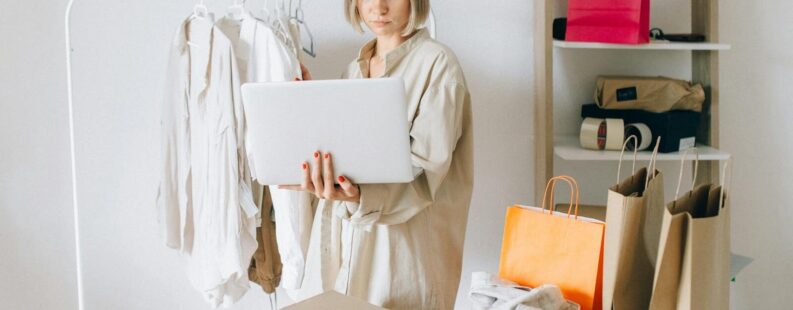 Woman using laptop for online shopping with clothes and bags around her, indoors.